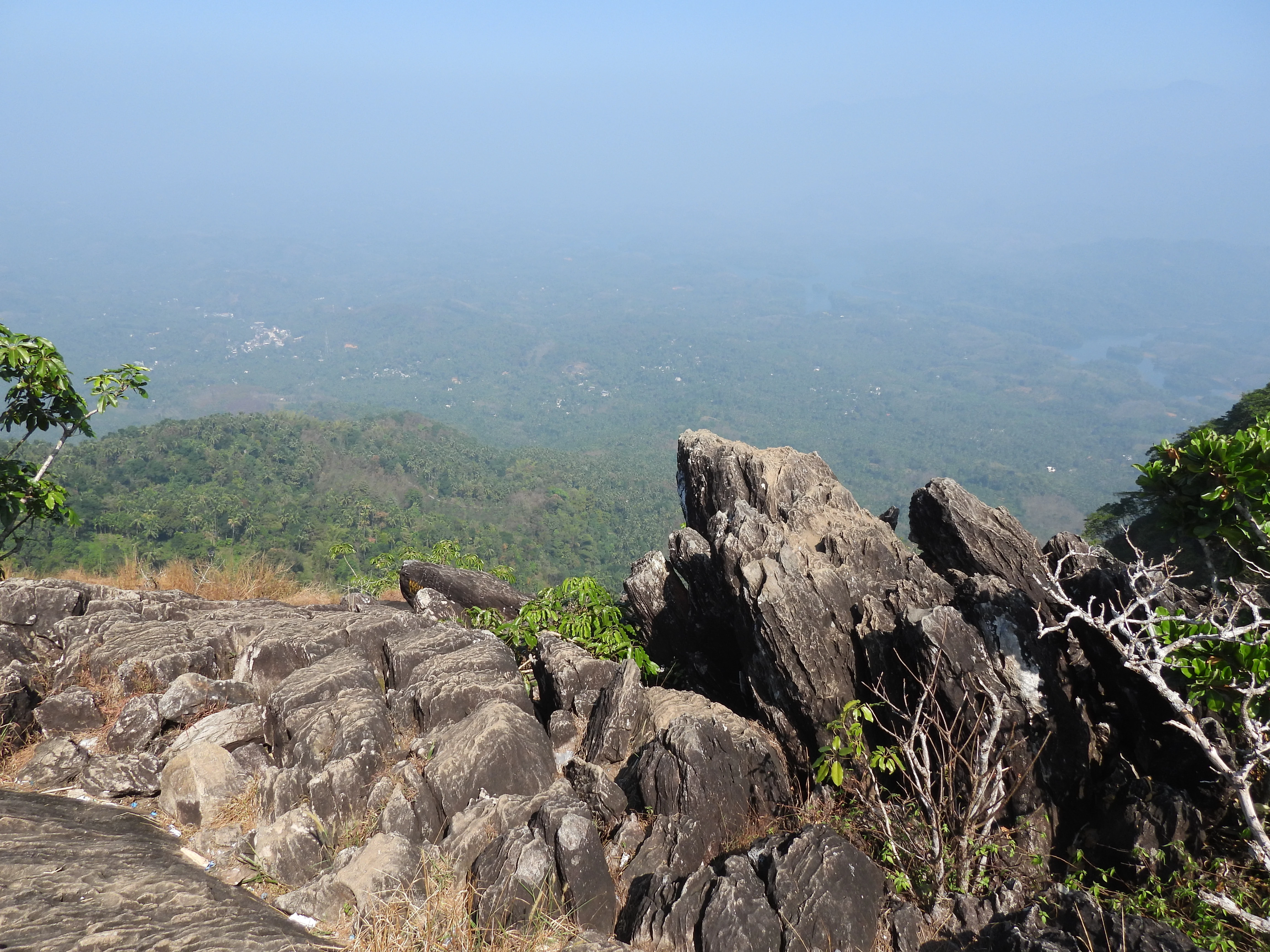 Jagged rock formations at a mountain viewpoint overlooking a vast green valley, forests, and distant hills under a clear blue sky.